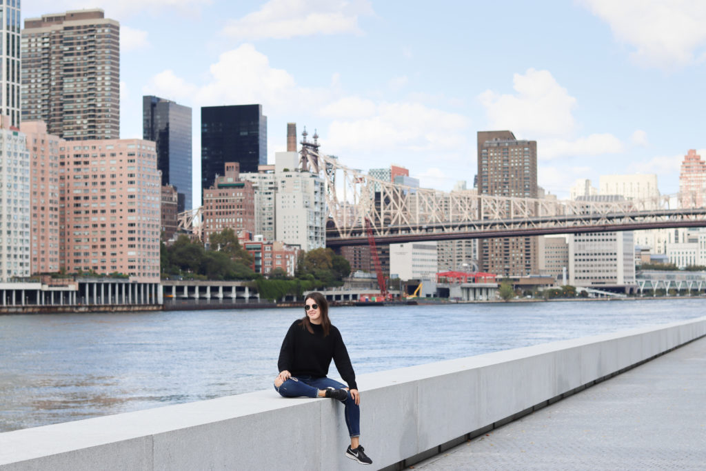 A woman sitting on a ledge with the NYC skyline and Ed Koch bridge in the background.