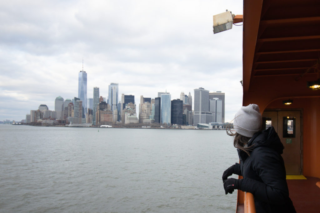 A woman standing on the Staten Island Ferry with the Manhattan skyline view in the background.