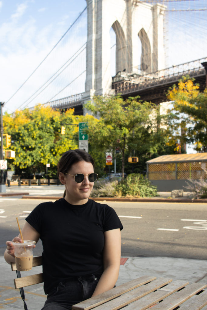 A women sitting with an iced coffee in front of the Brooklyn Bridge in Dumbo Brooklyn.