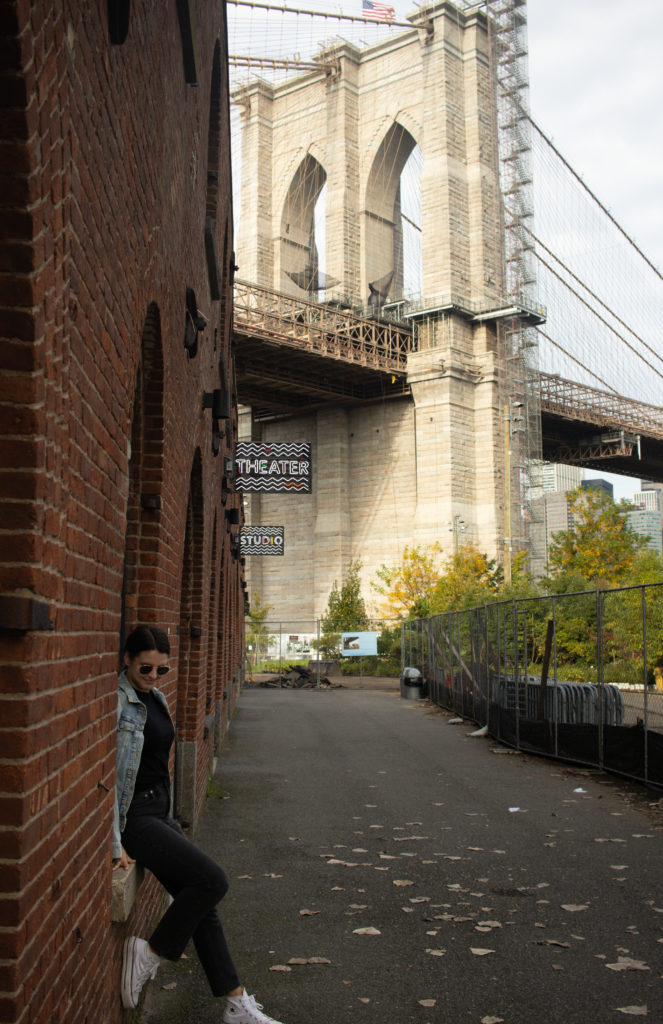 A women sitting along an industrial red-brick building in Dumbo Brooklyn with the Brooklyn Bridge in the background.